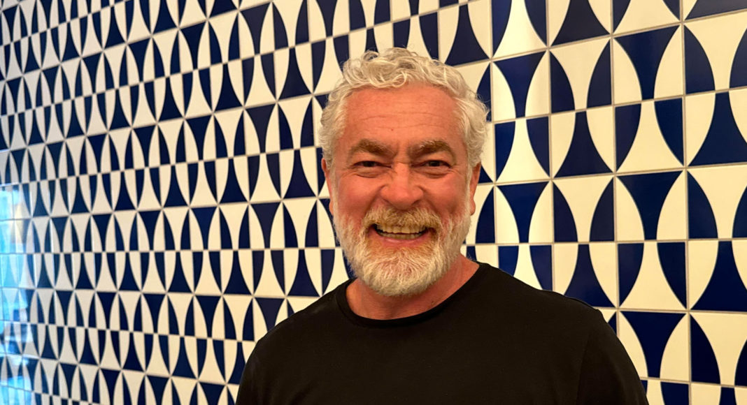 Man with a beard wearing a black t-shirt and standing in front of a a wall with a geometric black pattern