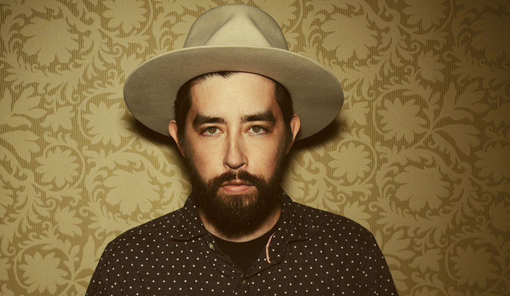 Bearded man wearing a hat and standing against a wallpaper background