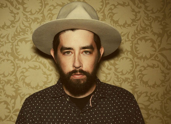Bearded man wearing a hat and standing against a wallpaper background