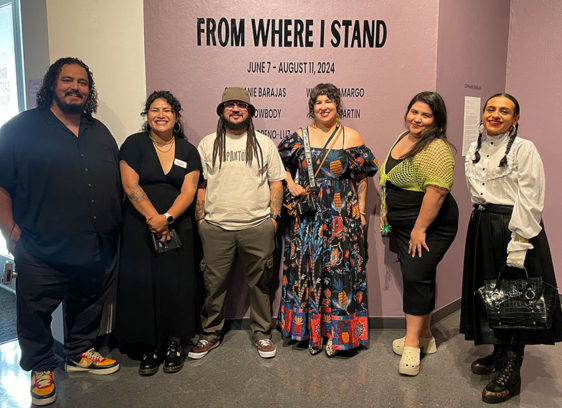 Six people in a gallery in front of a sign that says "From Where I Stand"