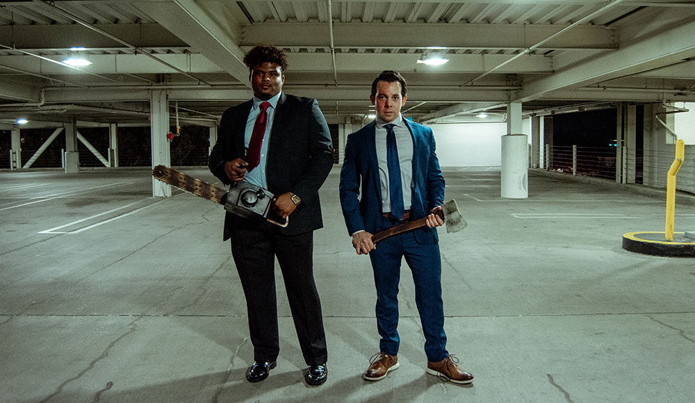 Two men in suits in a garage. The black man has a chainsaw; the white man has a hatchet