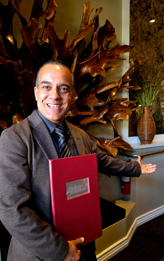 Man standing in front of a wooden sculpture, holding a menu and extending his arm in welcome