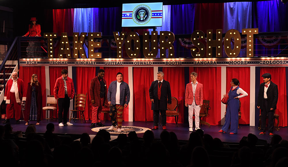 Nine performers on a stage, each standing under a sign with their character's name