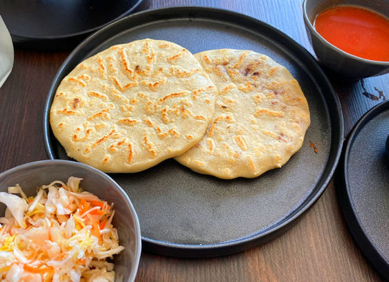 Two pupusas on a plate with bowls of cabbage and red salsa on the side