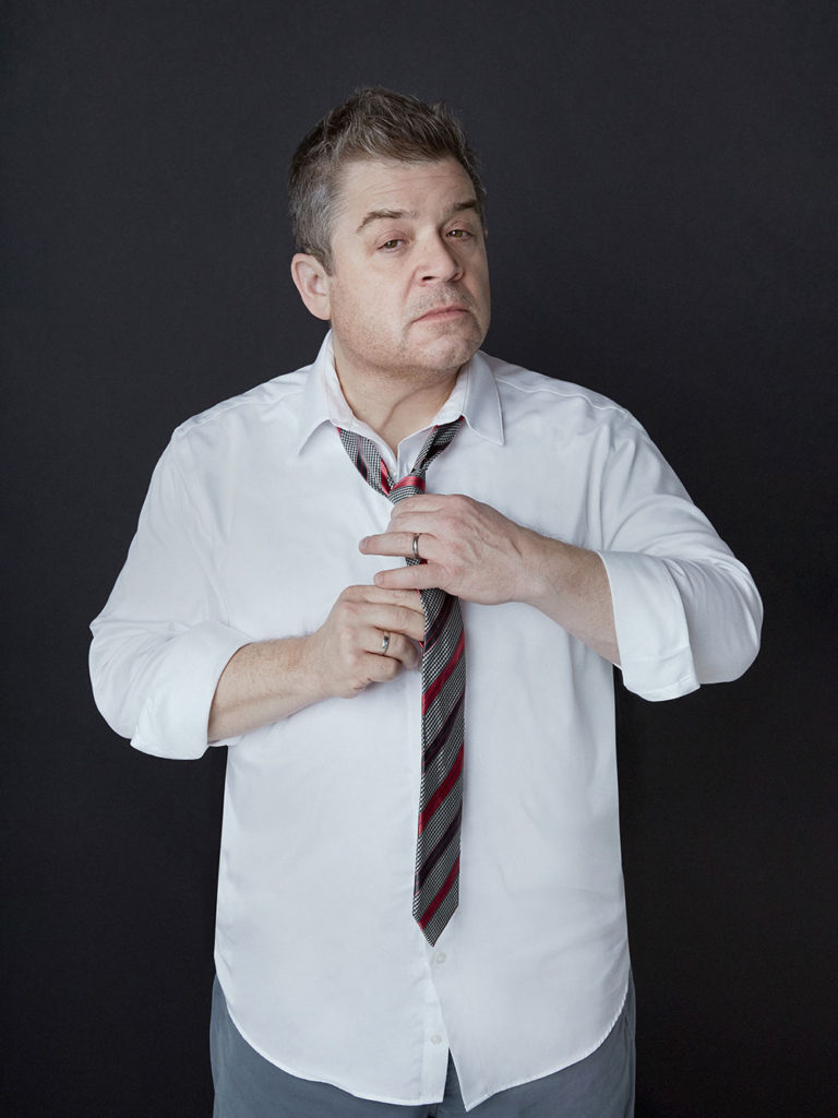 Patton Oswalt in shirtsleeves putting a tie on while grimacing at the camera.