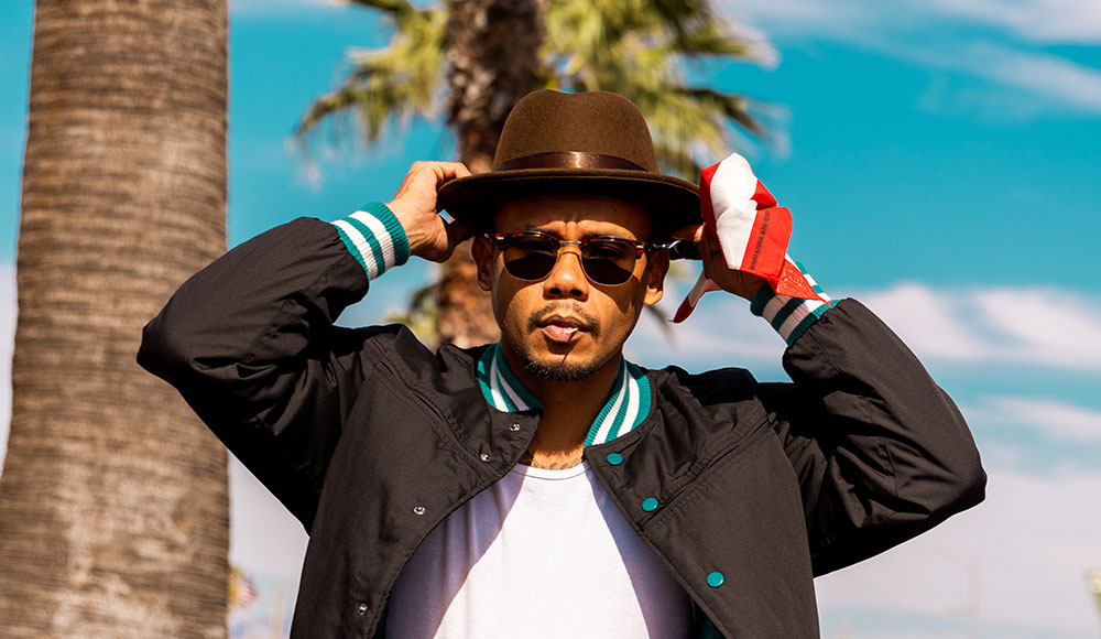 Man in sunglasses, his hands touching his hat, standing by palm trees