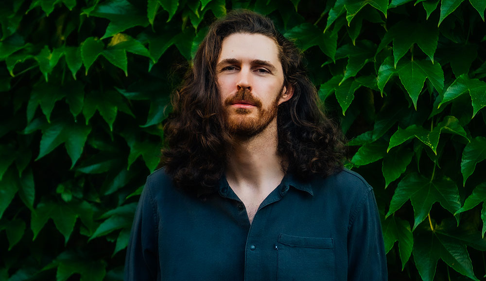 Pale man with dark long hair and beard in front of greenery