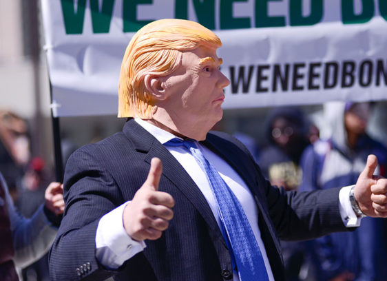 Man dressed in a suit wearing a Donald Trump mask at a rally