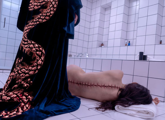 Woman with stitches along the length of her spine crumpled on a bathroom floor with a caped figure looming over her