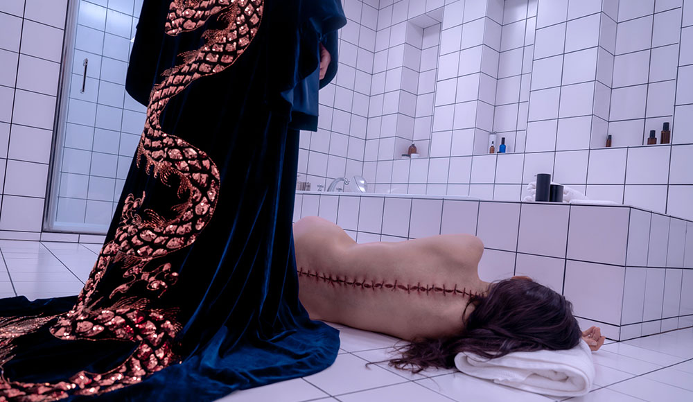 Woman with stitches along the length of her spine crumpled on a bathroom floor with a caped figure looming over her