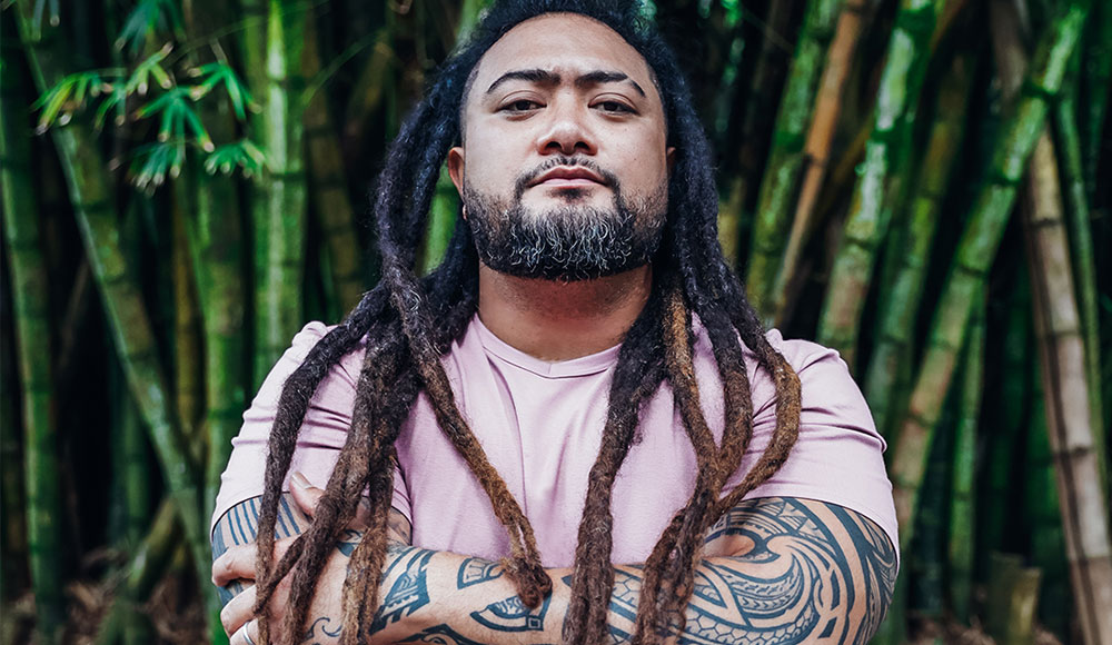 Photo of man with his arms folded, with tattooed arms and dreadlocks