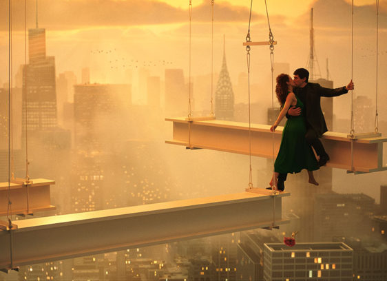 Man and woman hugging while standing on girders high above a cityscape