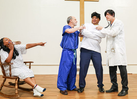 Four actors in a scene set in an insane asylum with one man in a straitjacket