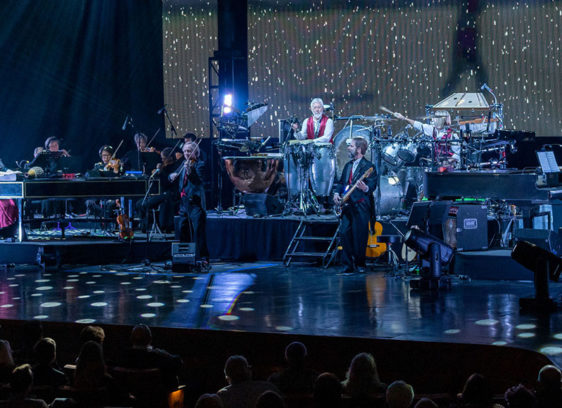 Large ensemble of musicians playing on a stage with holiday lights