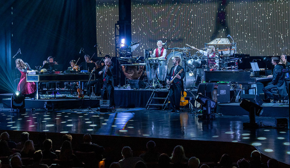 Large ensemble of musicians playing on a stage with holiday lights