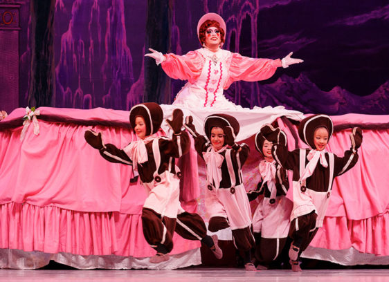 Performer in a pink dress with a huge skirt from which aa group of children in costumes are emerging