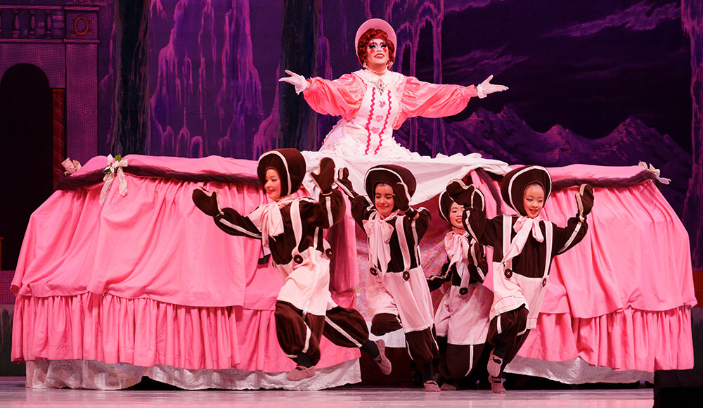 Performer in a pink dress with a huge skirt from which aa group of children in costumes are emerging