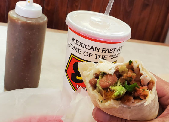 Hand holding a burrito with a drink and hot sauce dispenser also on the table