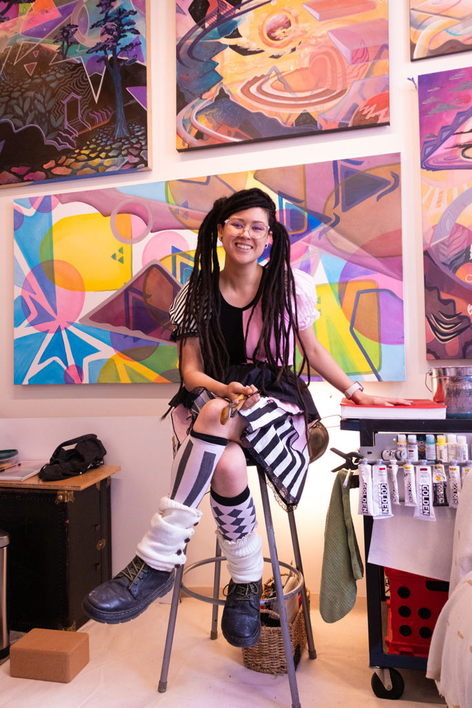 Woman sitting on a stool wearing whimsical clothing, paintings on the wall behind her.