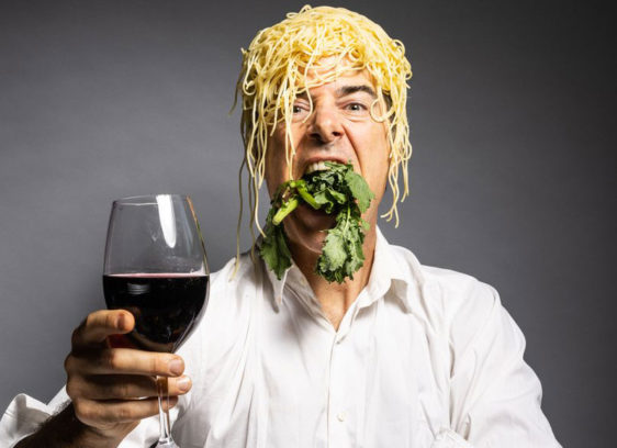 Man with a pile of noodles on his head toasting the camera with a glass of wine