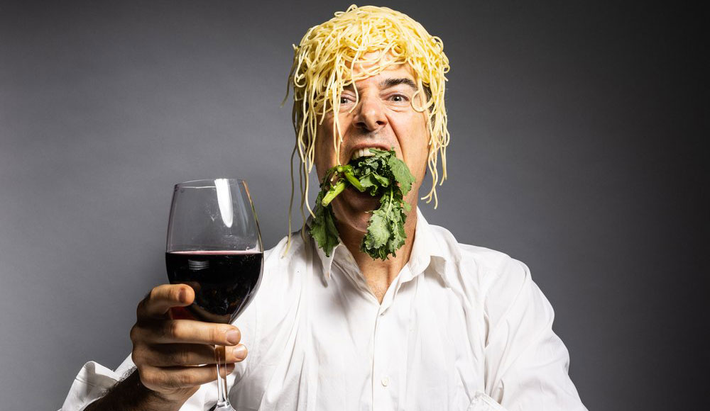 Man with a pile of noodles on his head toasting the camera with a glass of wine